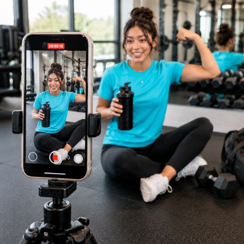 Young woman filming fitness content on a smartphone mounted to a tripod in a modern gym, sitting on the floor with a water bottle while flexing her arm in front of dumbbells and mirrors.